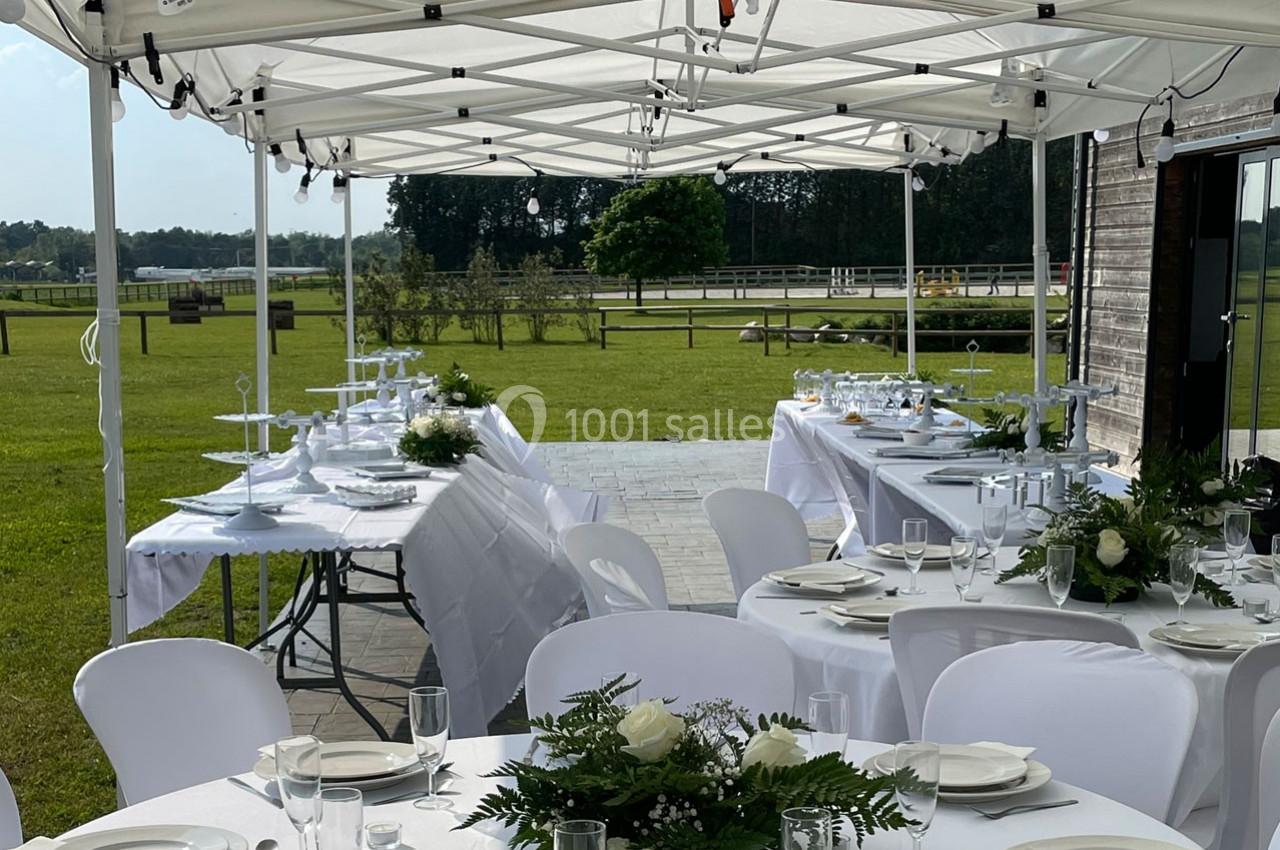 Tables dressées avec nappes blanches et décorations florales sous des tonnelles, dans un espace extérieur verdoyant.