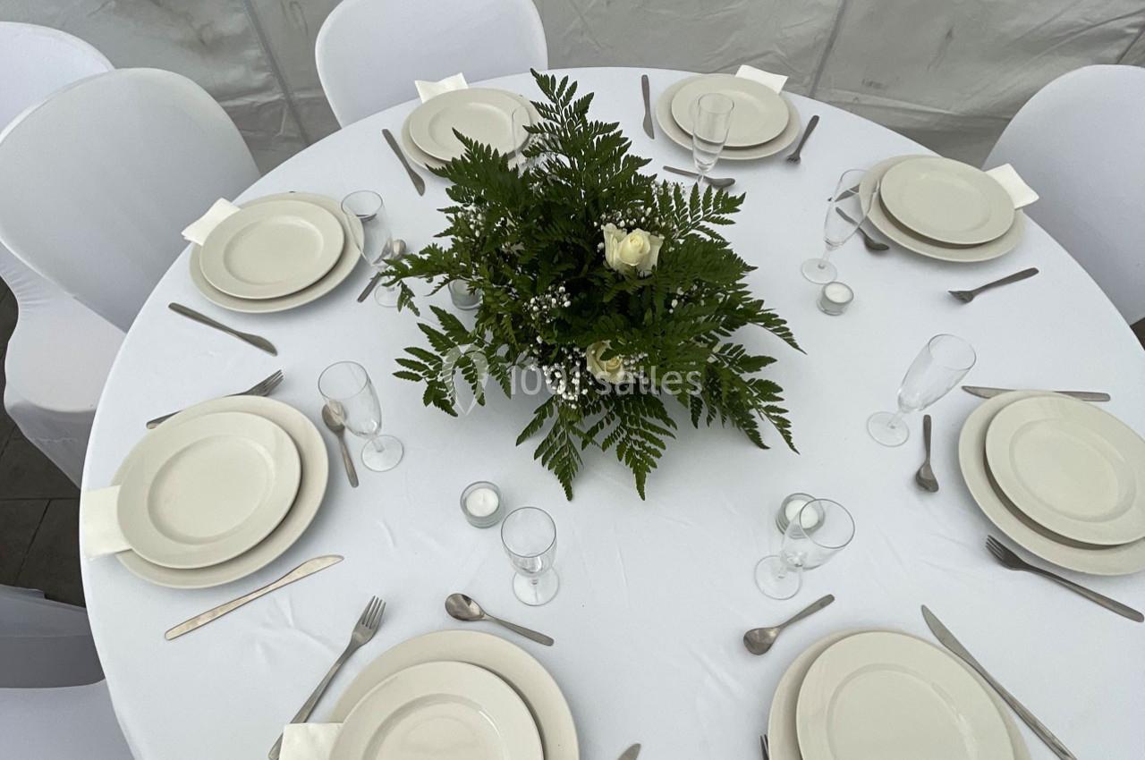 Table ronde dressée avec une nappe blanche, vaisselle blanche, couverts en métal et un centre de table floral.