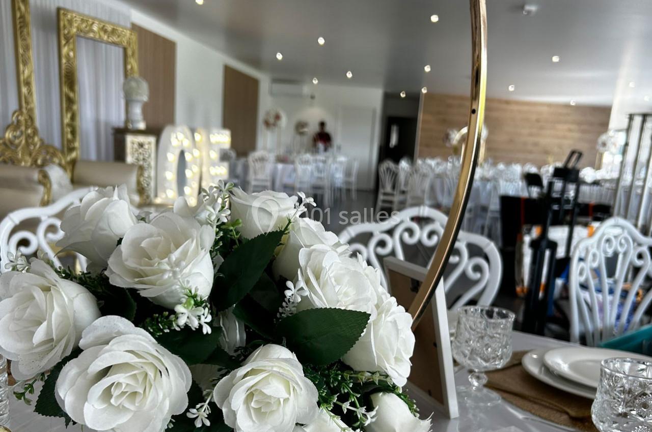 Centre de table avec des roses blanches dans une salle de réception élégante, décorée de chaises blanches et de cadres dorés.