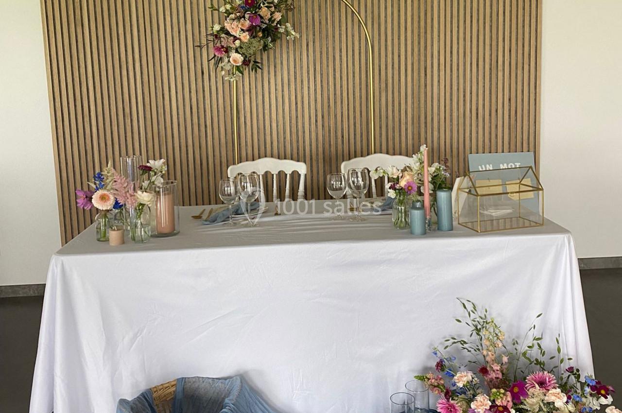 Table décorée pour un mariage avec nappes blanches, fleurs colorées, verrerie et arche florale en arrière-plan.