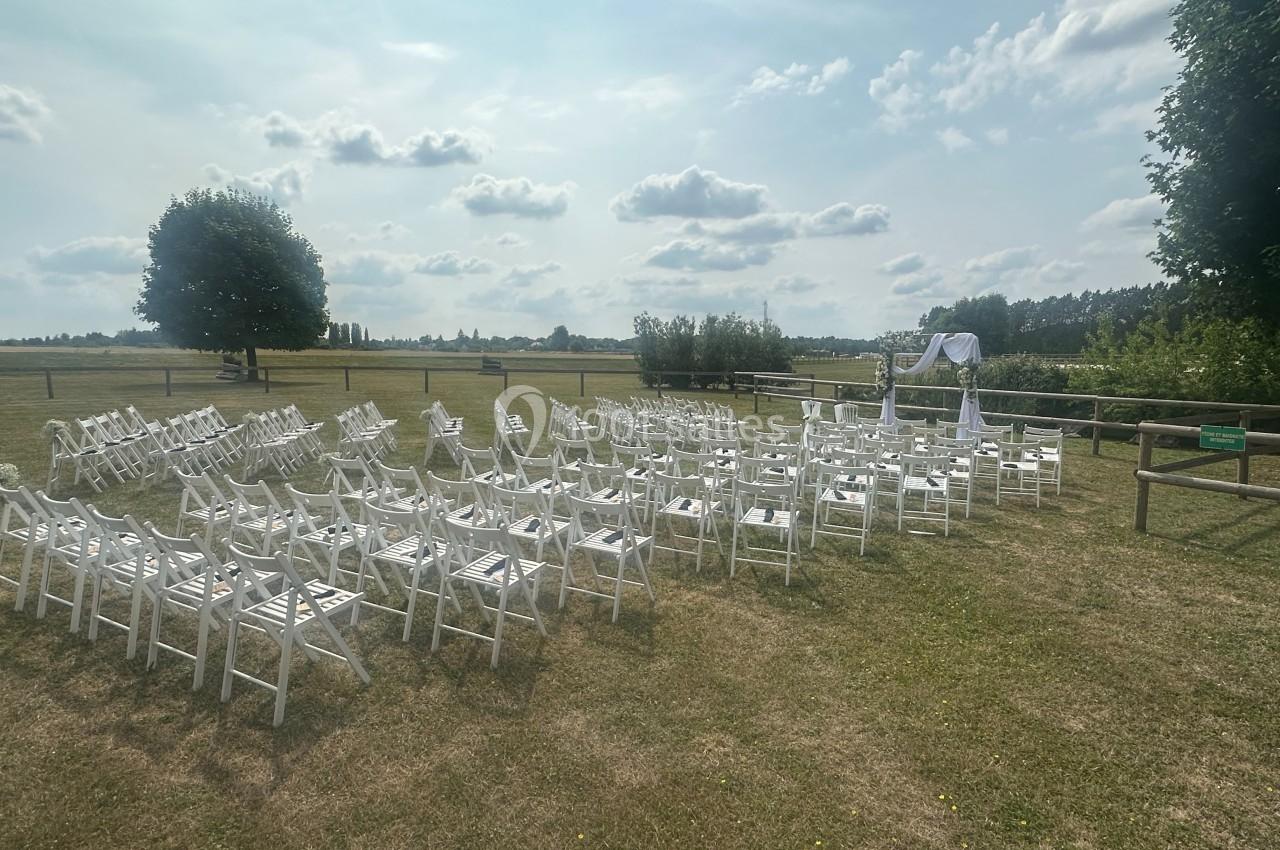 Chaises blanches disposées en rangées sur une pelouse, face à une arche décorée, dans un cadre extérieur champêtre.