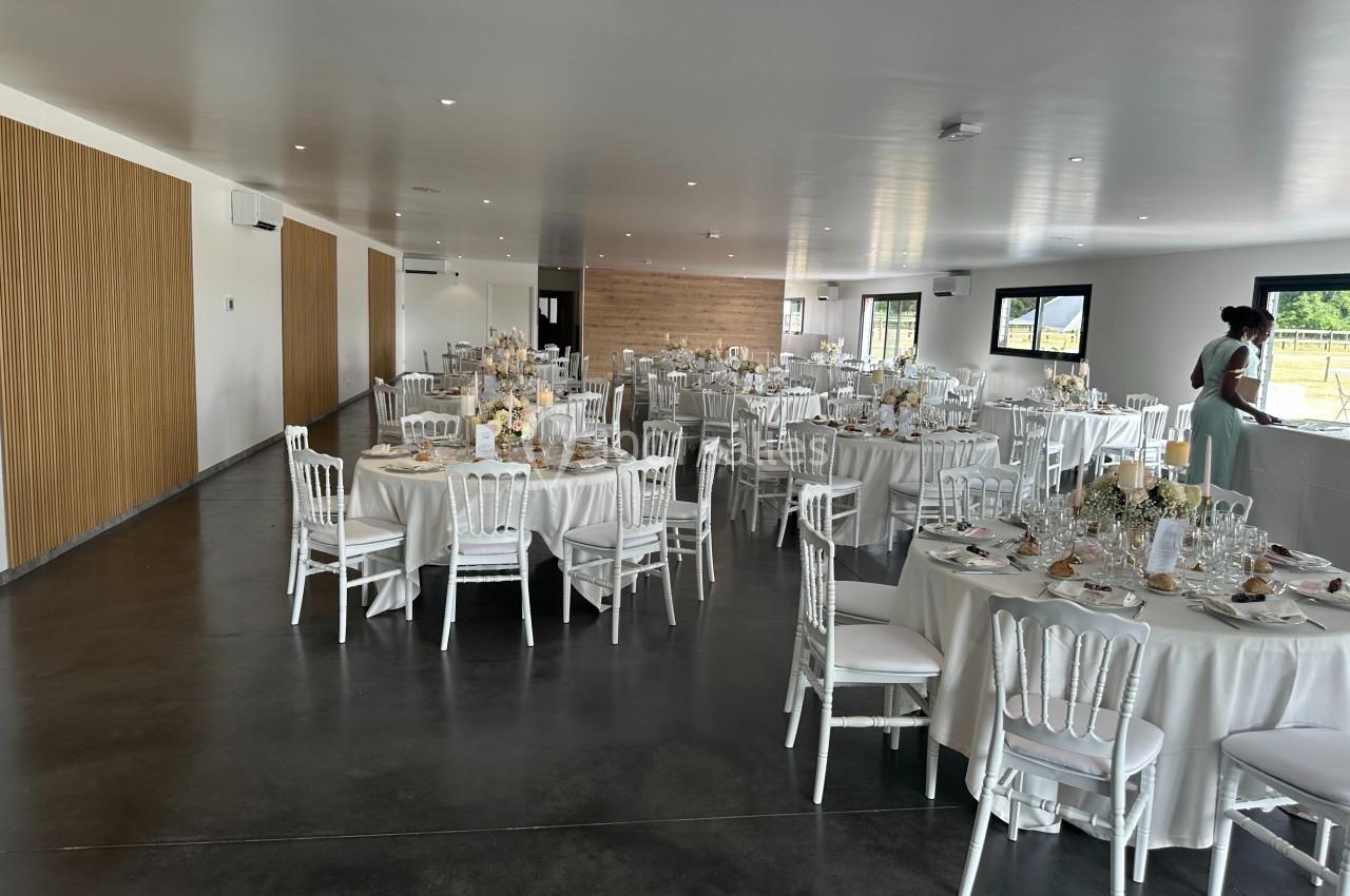 Salle de réception lumineuse avec tables rondes dressées, chaises blanches et décorations florales élégantes.
