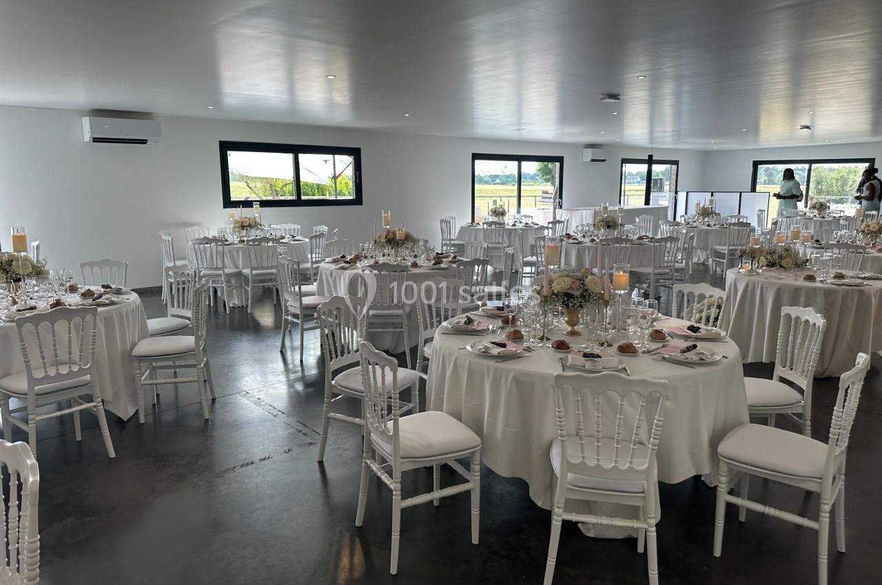 Salle de réception lumineuse avec tables rondes décorées de nappes blanches, fleurs et vaisselle élégante.