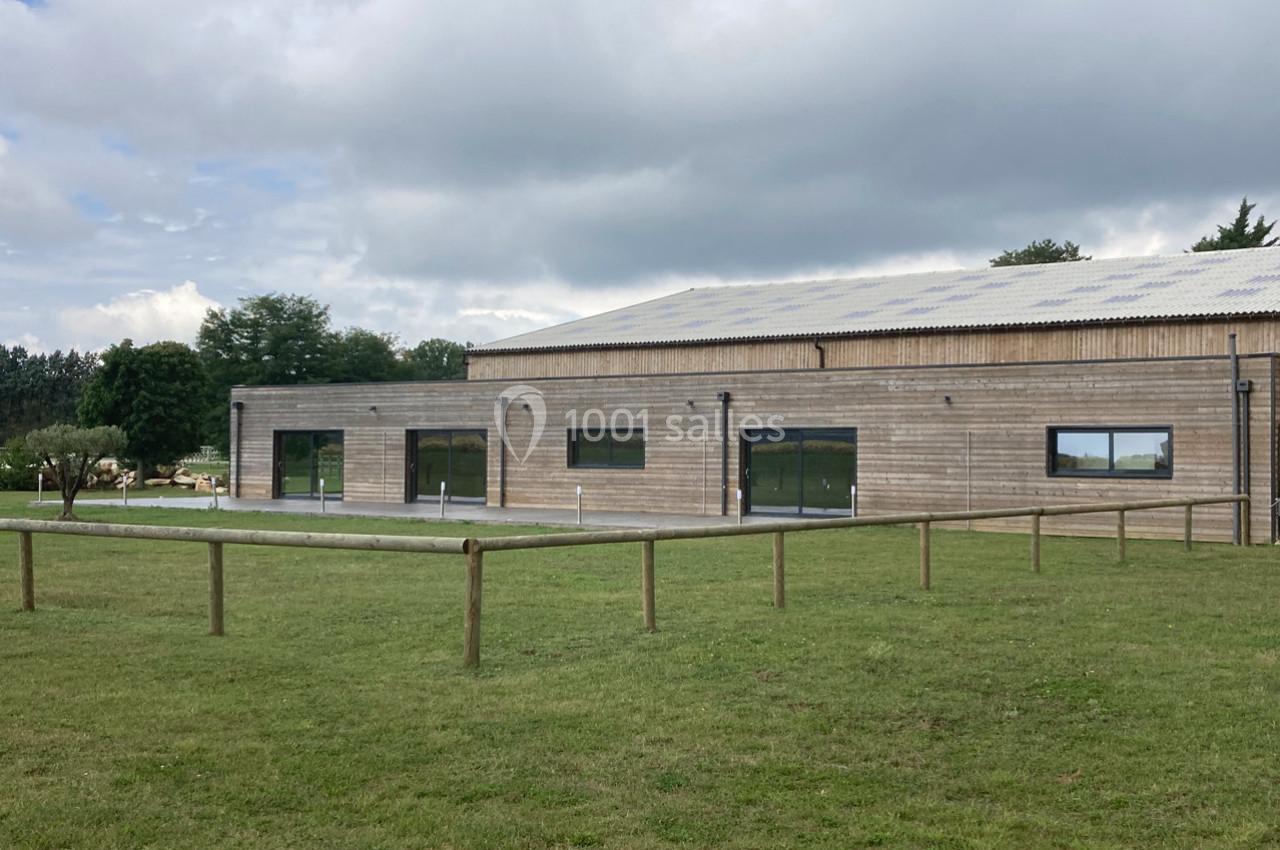 Bâtiment en bois avec de grandes baies vitrées, entouré d'une pelouse et d'une clôture en bois, sous un ciel nuageux.