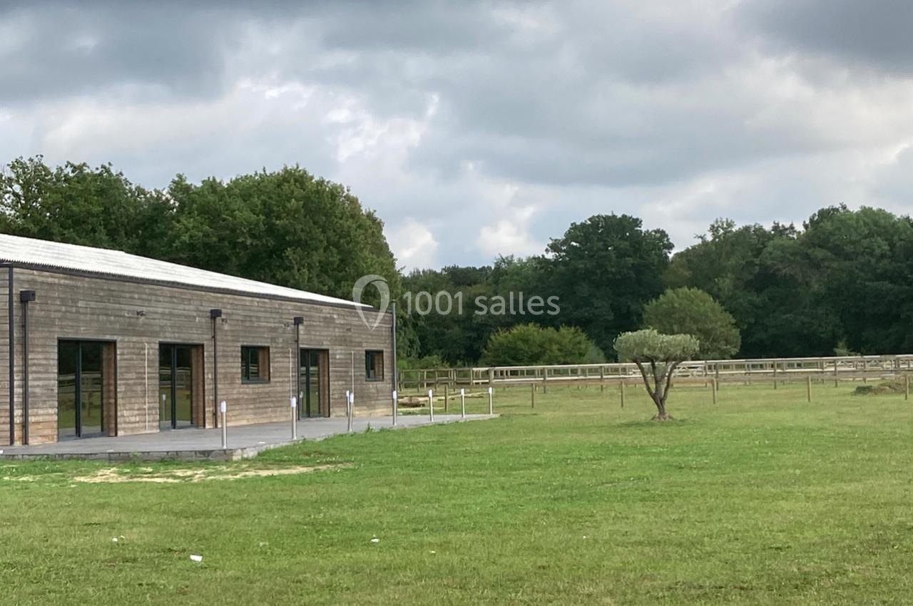 Bâtiment en bois moderne entouré d'une pelouse, avec des arbres et une clôture en arrière-plan sous un ciel nuageux.