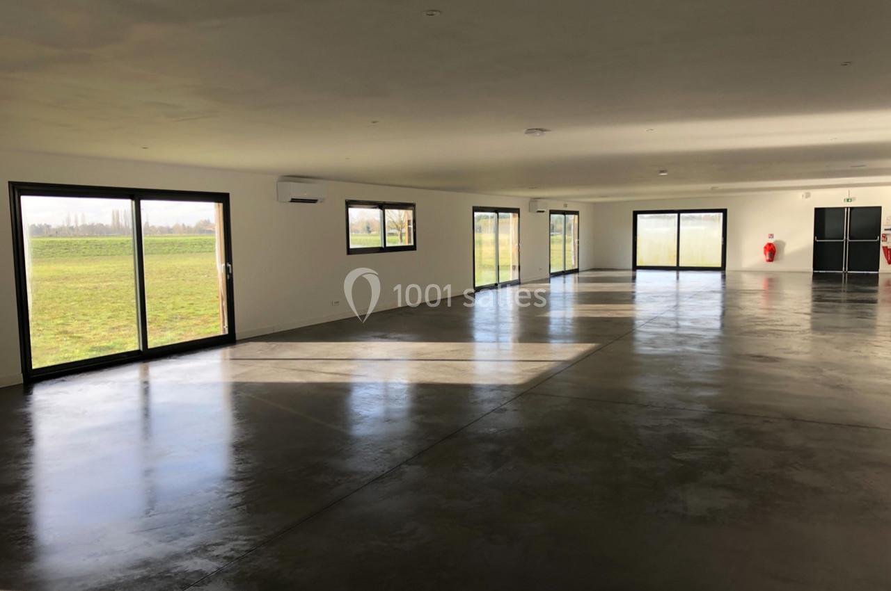 Salle vide avec sol en béton poli, grandes baies vitrées laissant entrer la lumière naturelle et vue sur un espace vert.