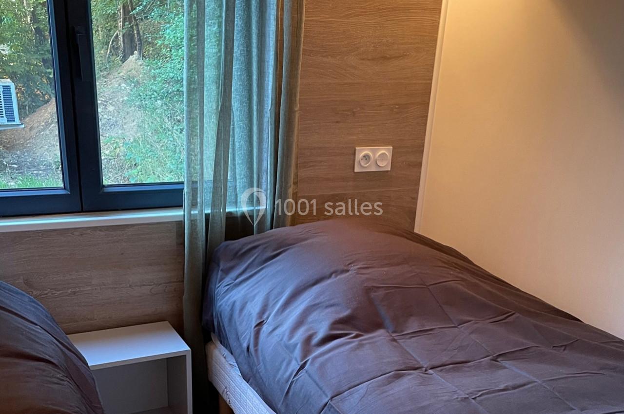 Deux lits simples avec draps gris dans une chambre en bois, éclairée par une fenêtre avec vue sur des arbres.