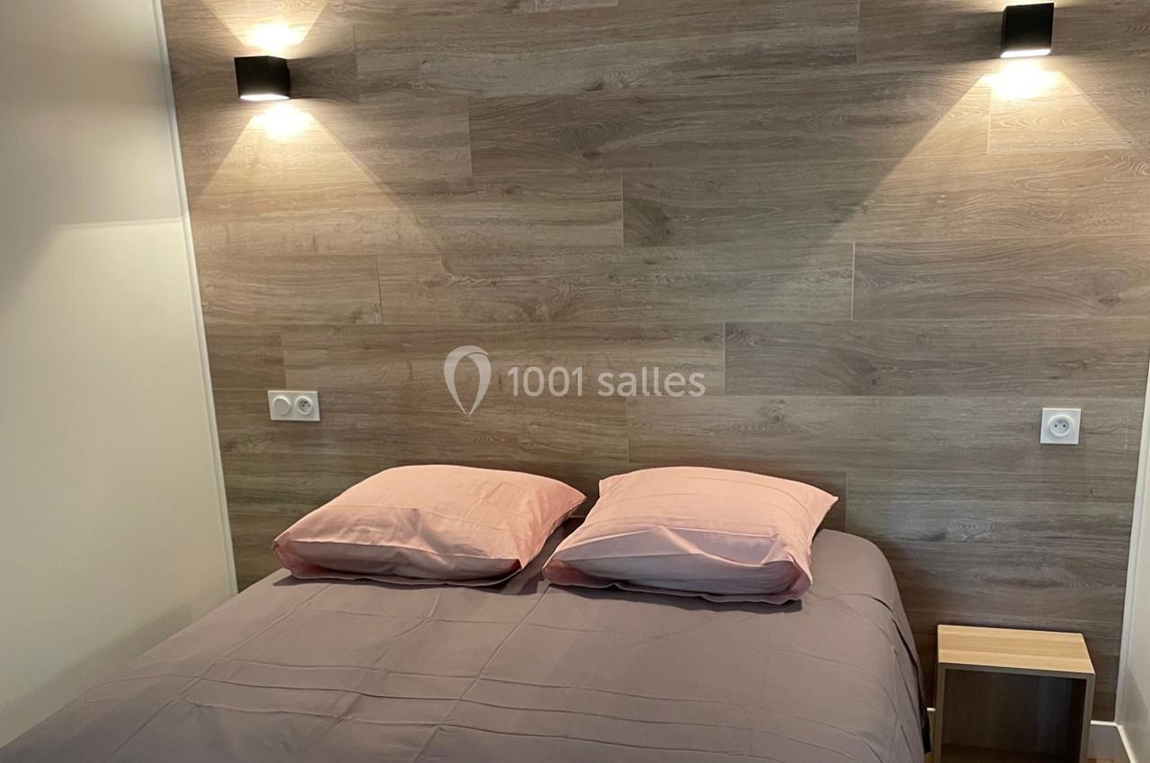 Lit double avec draps gris et oreillers roses, encadré par deux appliques murales sur un mur en bois clair.