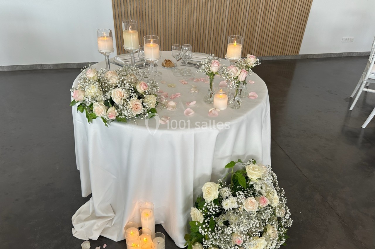 Table ronde décorée pour un mariage avec nappes blanches, bougies, pétales de rose et arrangements floraux.