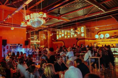 Salle lumineuse avec murs rouges, tables hautes en bois, tabourets métalliques et décorations murales colorées.