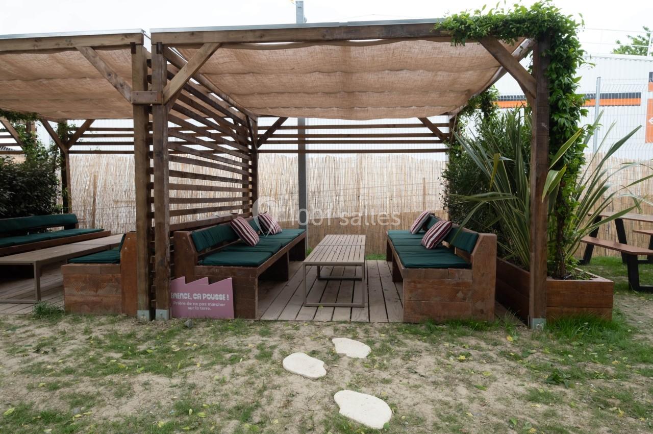 Espace extérieur avec pergolas en bois, banquettes vertes, table basse et végétation environnante.