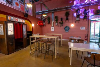 Salle lumineuse avec murs rouges, tables hautes en bois et métal, tabourets, affiches décoratives et luminaires modernes.
