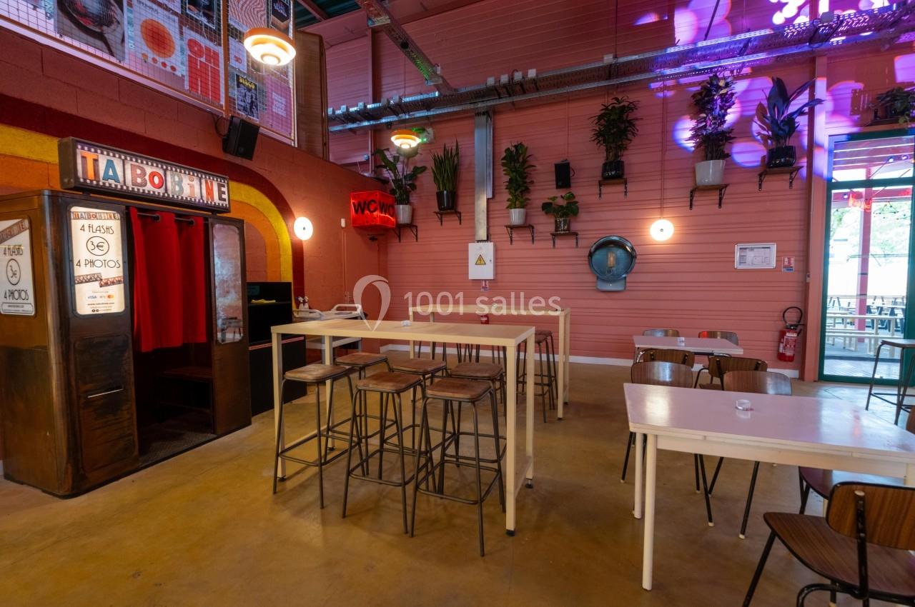 Espace intérieur coloré avec tables hautes, chaises, plantes murales et un photomaton rétro dans un coin.