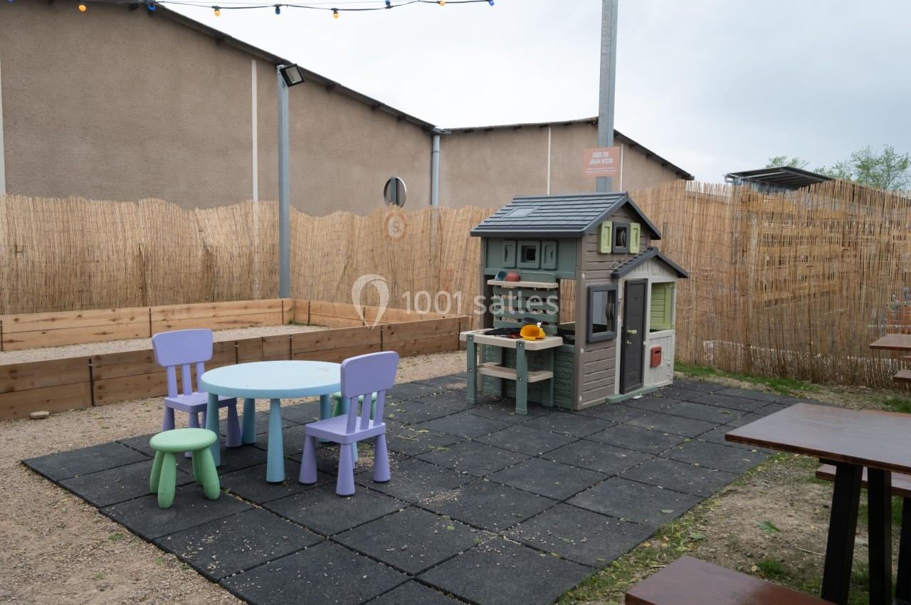 Aire de jeux extérieure avec une maisonnette en plastique, une table ronde et deux chaises colorées sur un sol en dalles.
