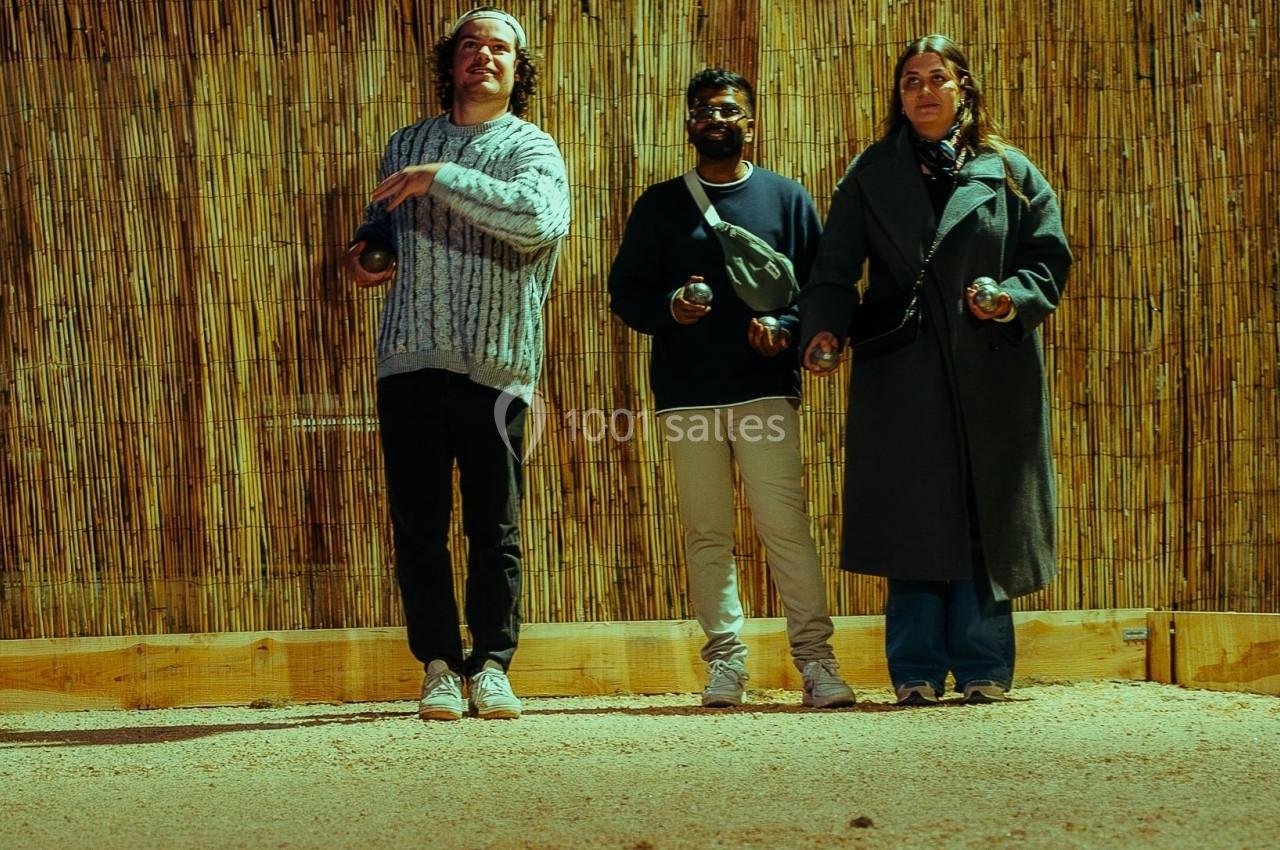 Trois personnes jouent à la pétanque sur un terrain en sable, avec des boules dispersées au sol.