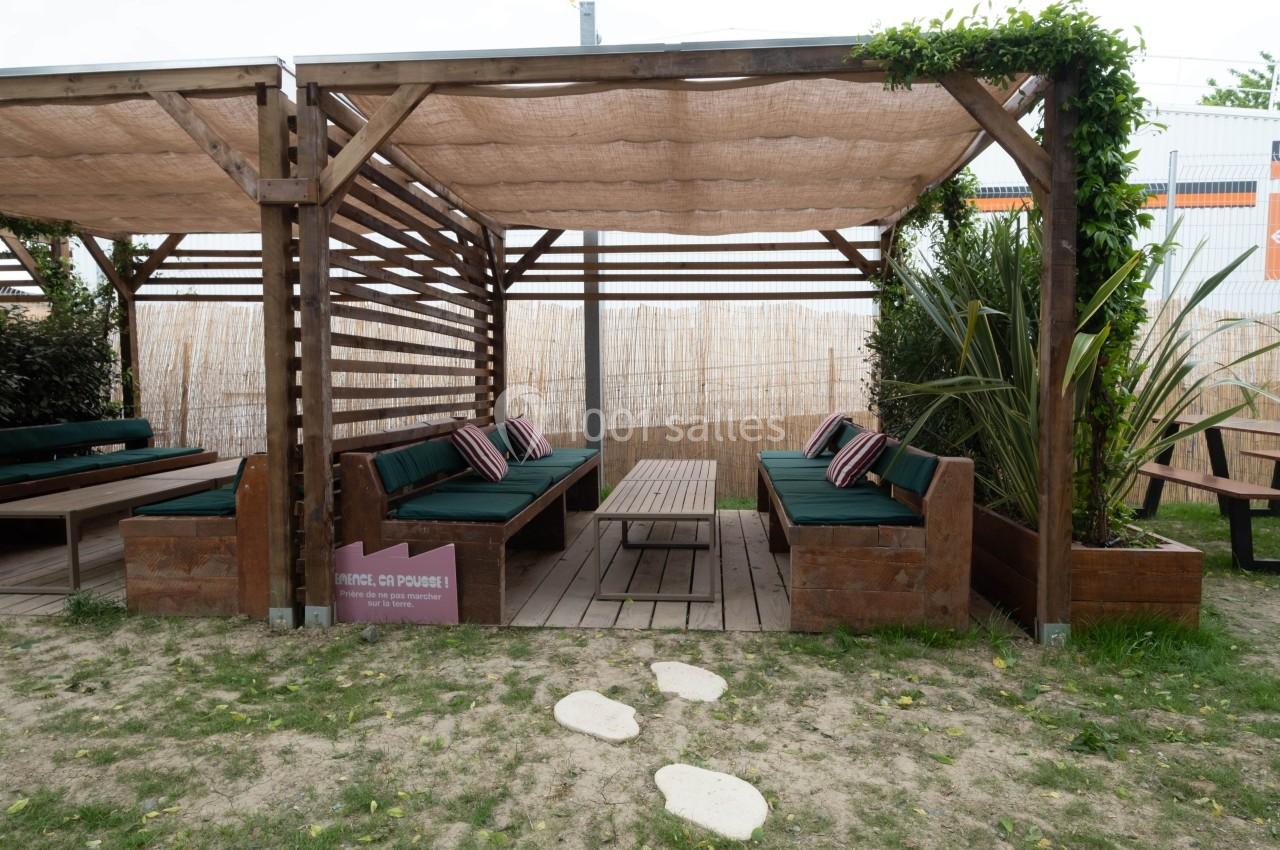 Espace extérieur avec pergolas en bois, banquettes vertes, table basse et végétation environnante.