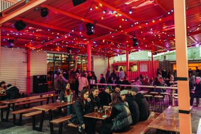 Salle lumineuse avec murs rouges, tables hautes en bois et métal, tabourets, affiches décoratives et luminaires modernes.