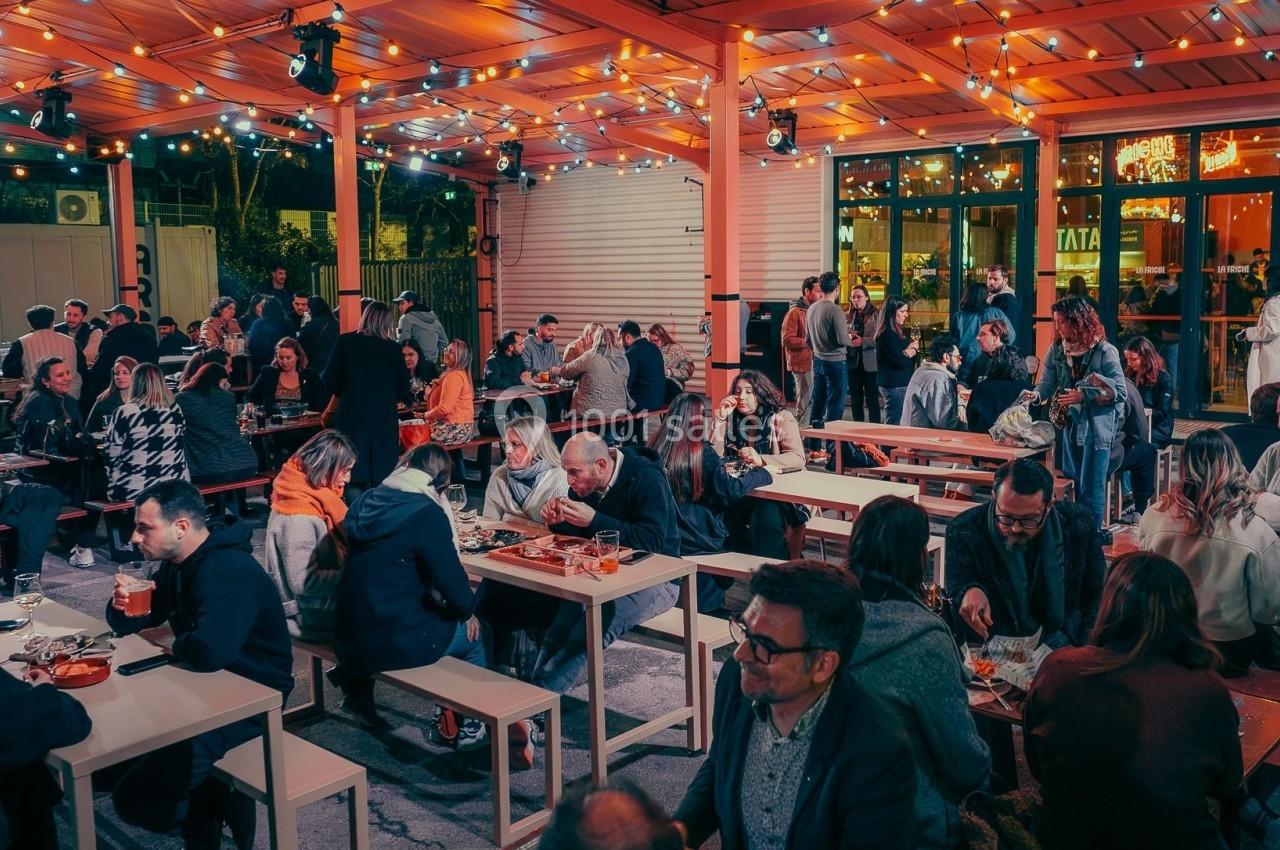 Terrasse animée en soirée avec des tables remplies de personnes partageant un repas sous des guirlandes lumineuses.