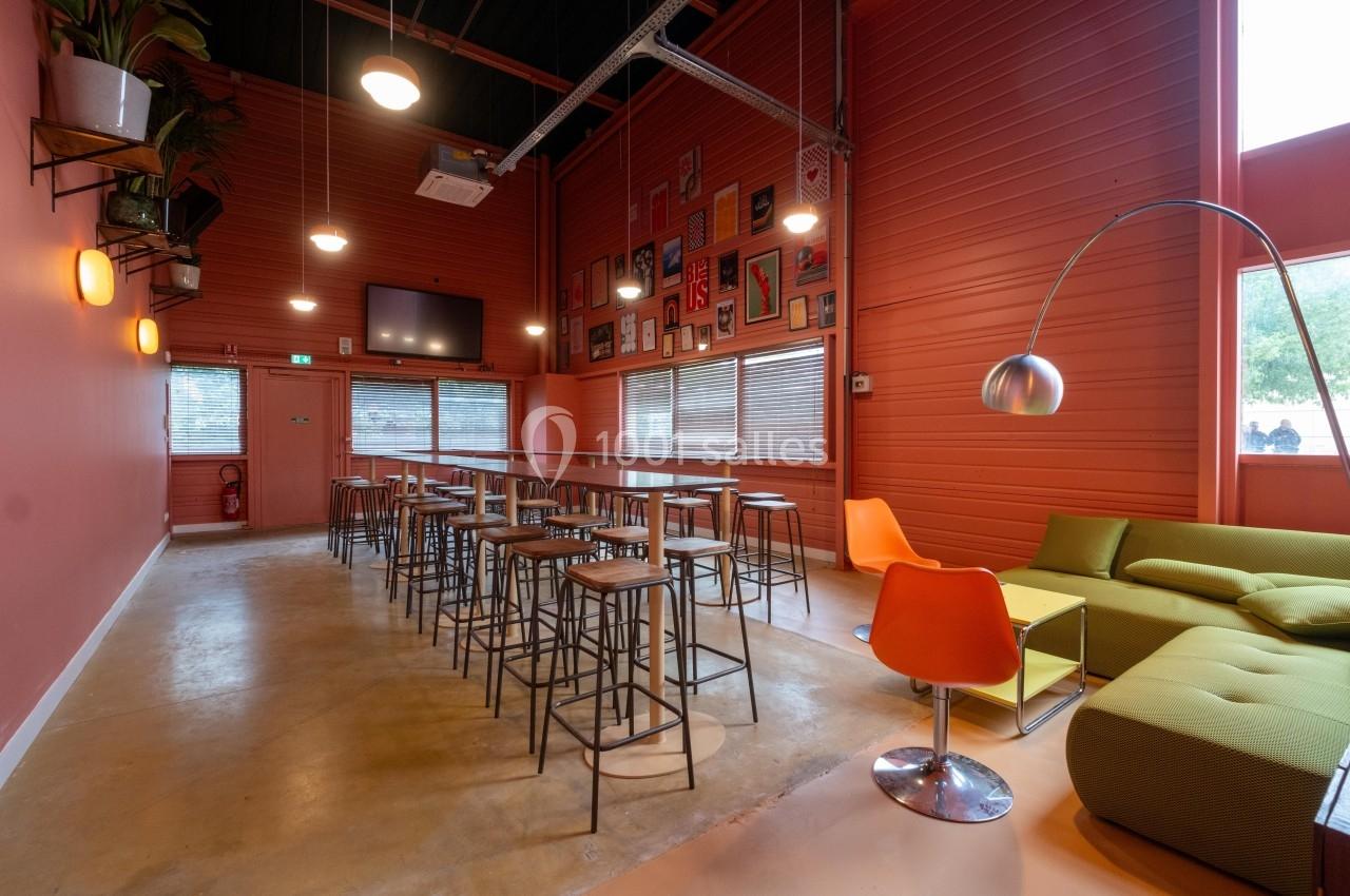 Salle lumineuse avec murs rouges, tables hautes, tabourets, canapé vert, chaise orange et décorations murales.