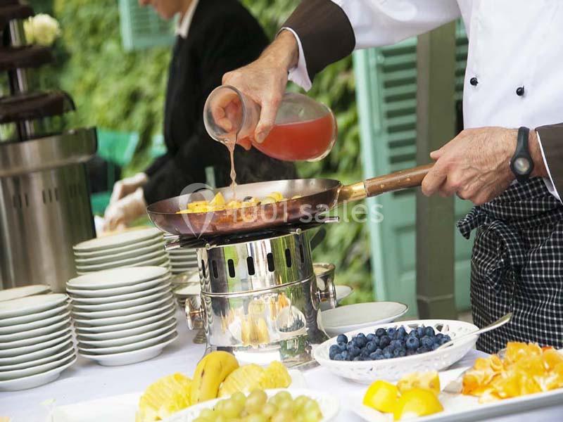 Un chef verse une sauce dans une poêle sur un réchaud, entouré de fruits frais et d'assiettes empilées.