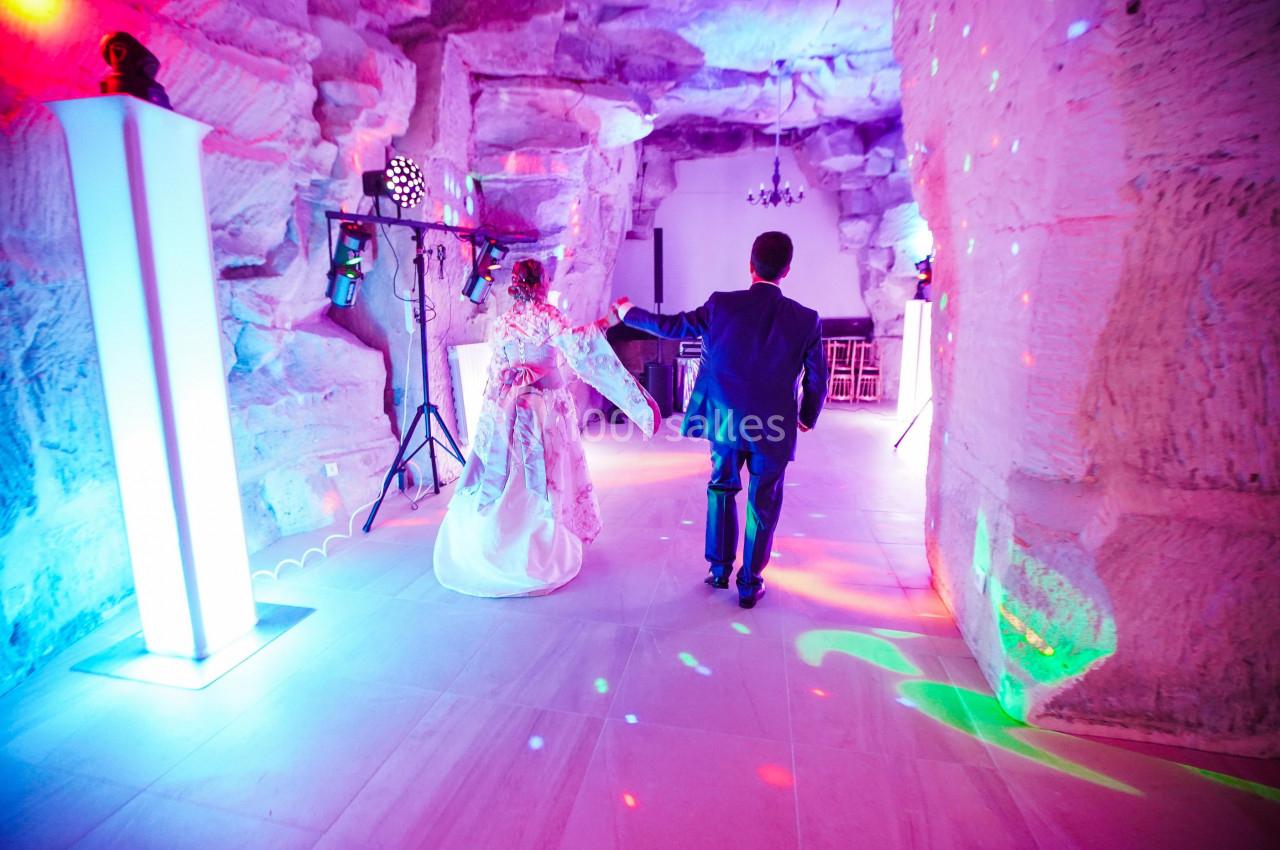 Un couple en tenue de mariage danse dans une salle aux murs de pierre éclairée par des lumières colorées.