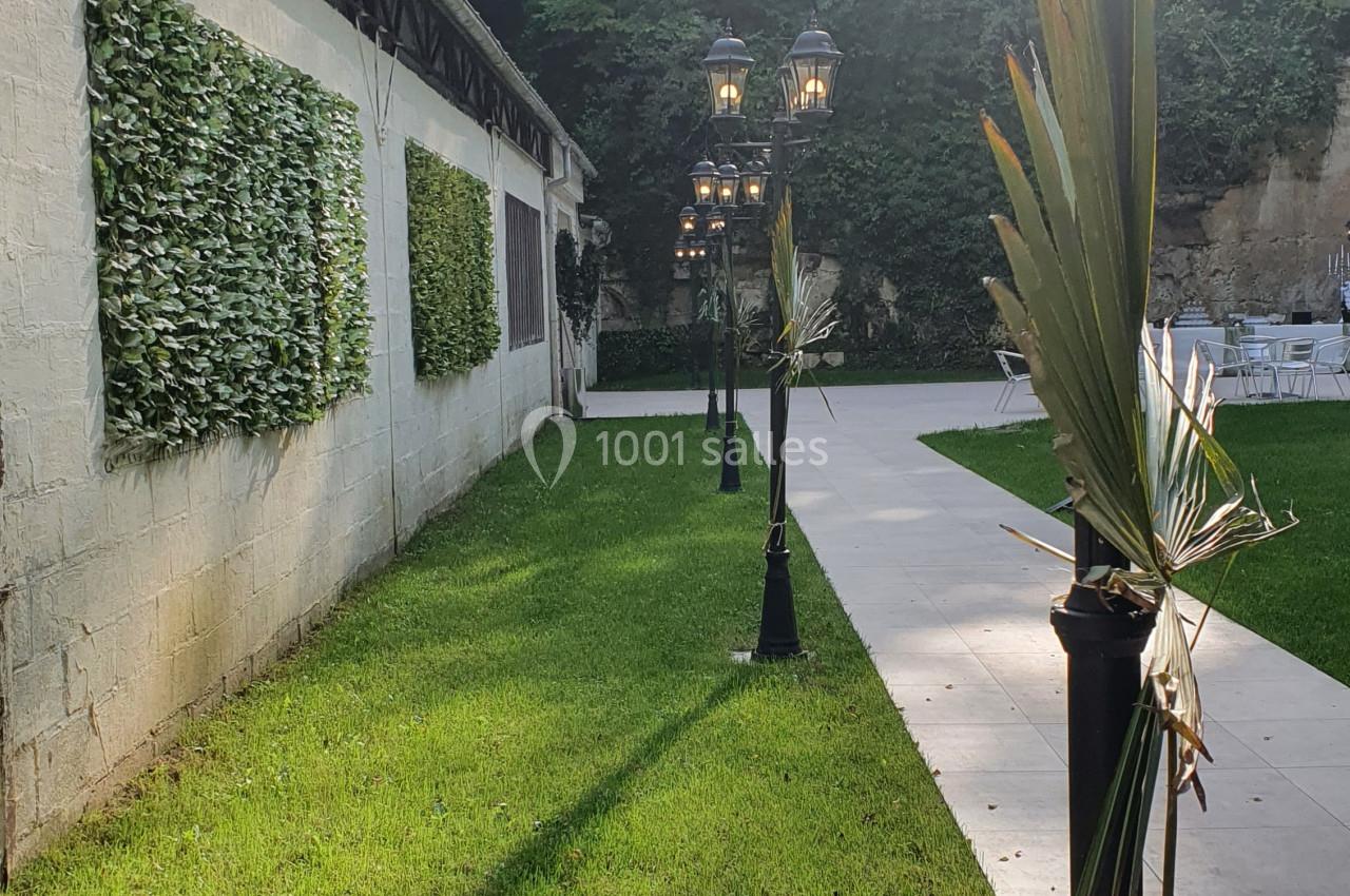 Allée bordée de lampadaires décorés de feuilles, longeant un mur végétalisé et un espace gazonné.