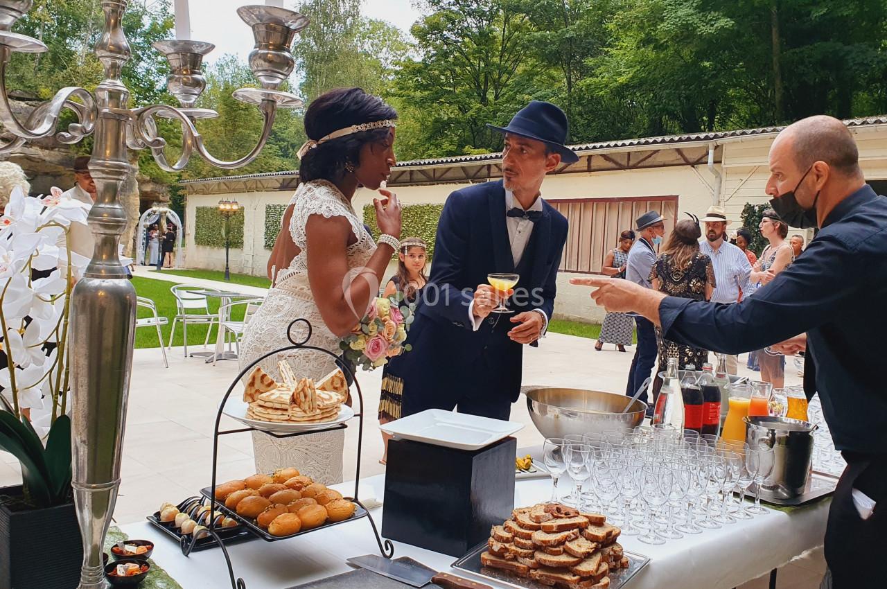 Un couple en tenue de mariage échange près d'un buffet en extérieur, entouré d'invités dans un cadre verdoyant.