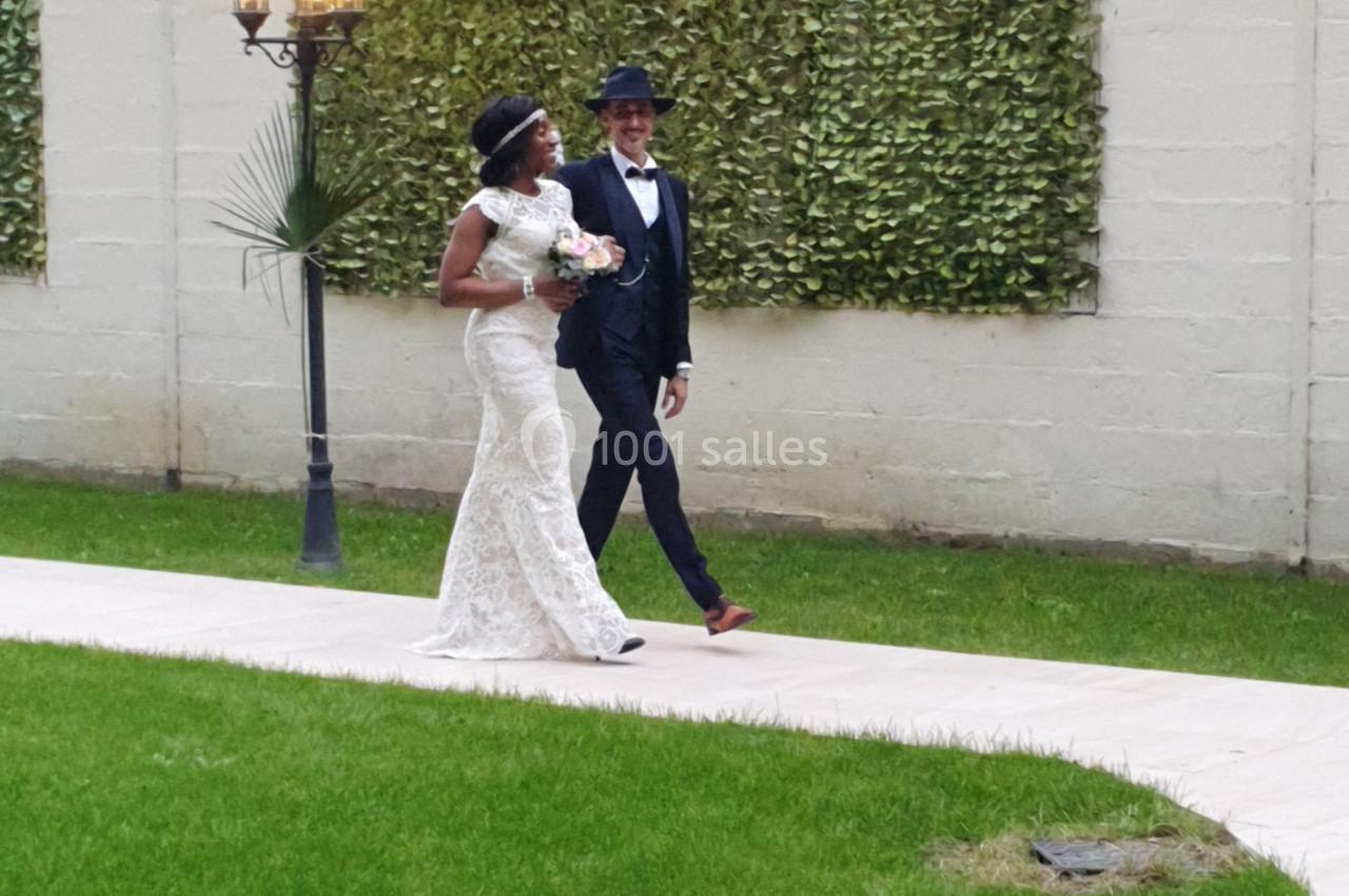 Un couple marche sur une allée pavée dans un jardin, la mariée en robe blanche tenant un bouquet.