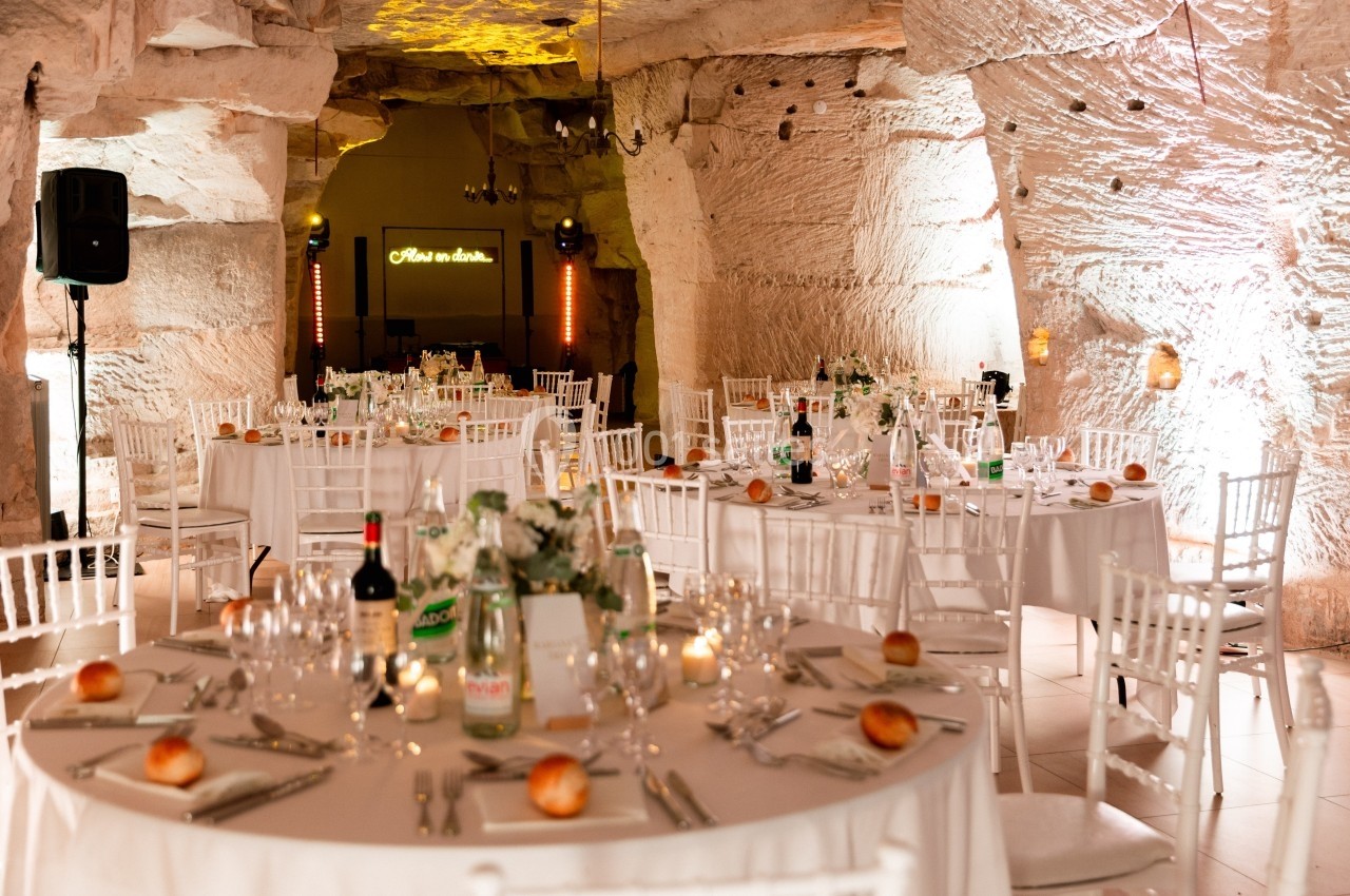 Salle de réception aménagée dans une grotte, avec tables rondes décorées, chaises blanches et éclairage chaleureux.