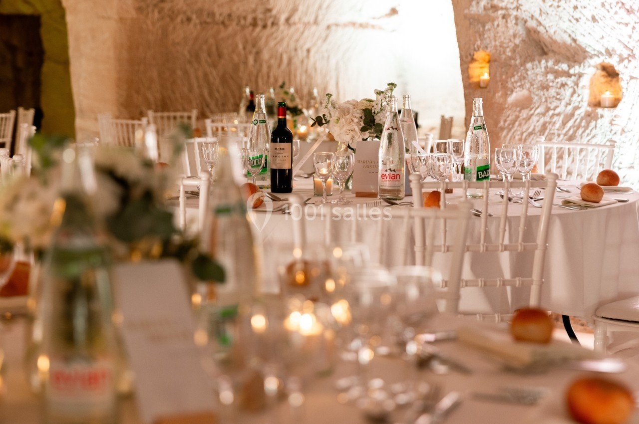 Tables élégamment dressées avec nappes blanches, chaises assorties, bouteilles d'eau et de vin, dans une salle éclairée.