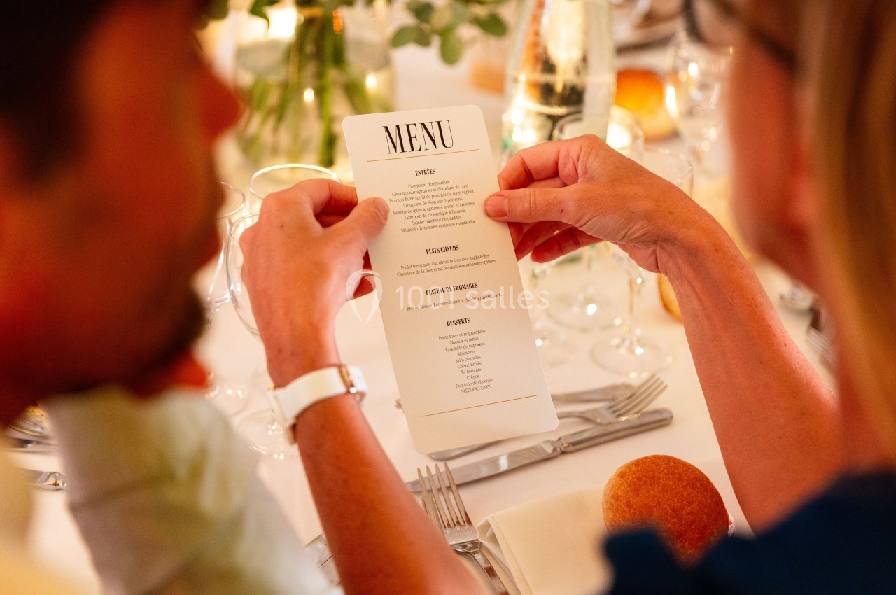 Deux personnes consultent un menu lors d'un repas, avec une table dressée et des éléments décoratifs en arrière-plan.