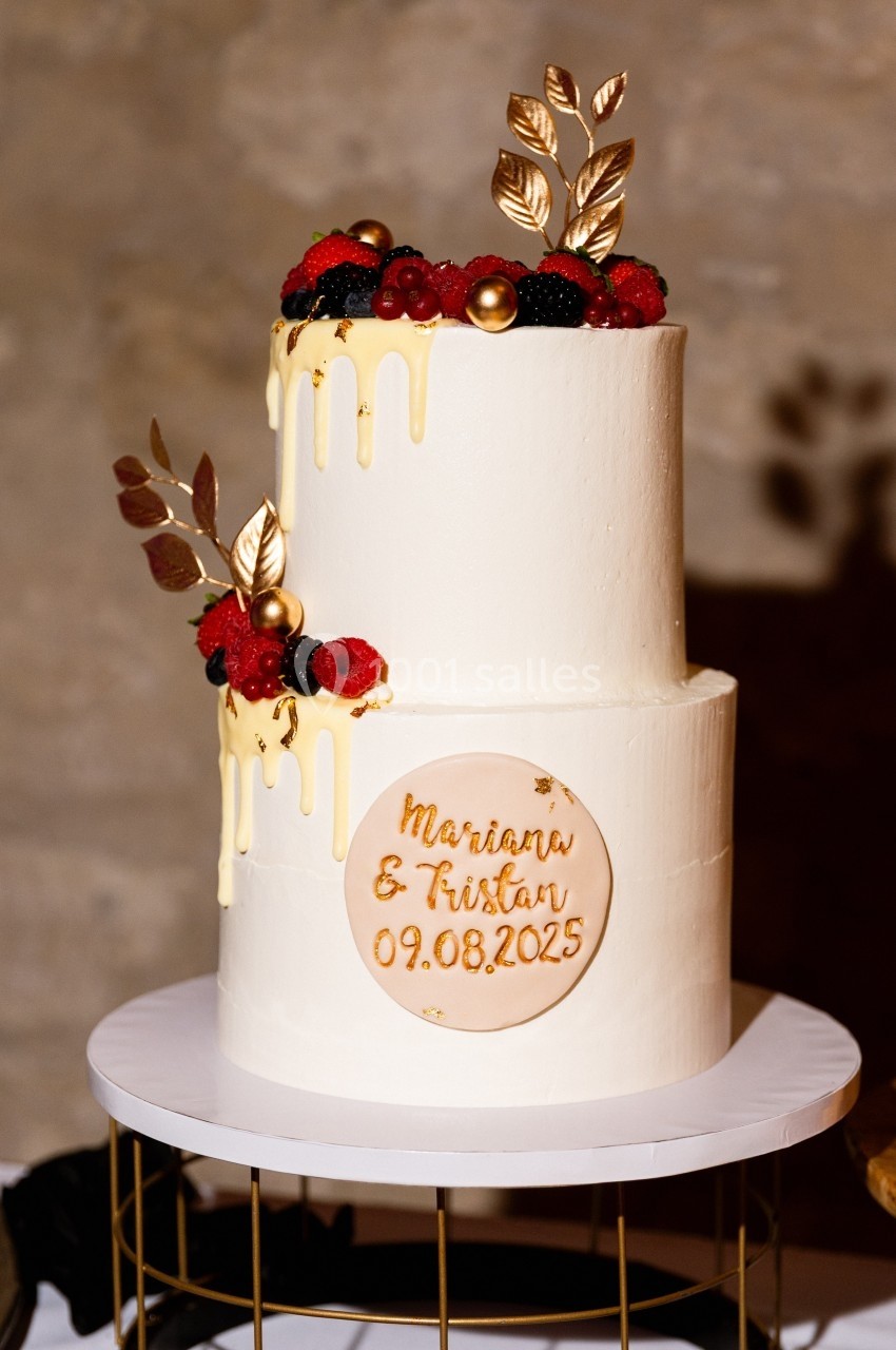Gâteau de mariage à deux étages décoré de fruits rouges, feuilles dorées et glaçage coulant.
