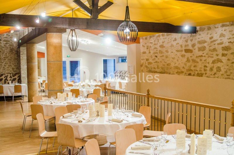 Salle de réception lumineuse avec tables rondes dressées, chaises en bois clair et murs en pierre apparente.