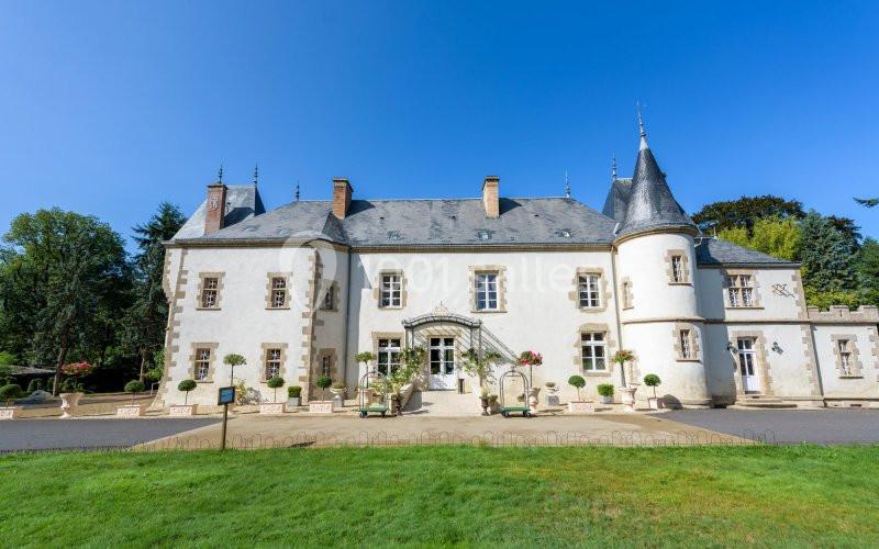 Façade d'un château ancien avec tours, toit en ardoise et jardin à l'avant sous un ciel bleu.