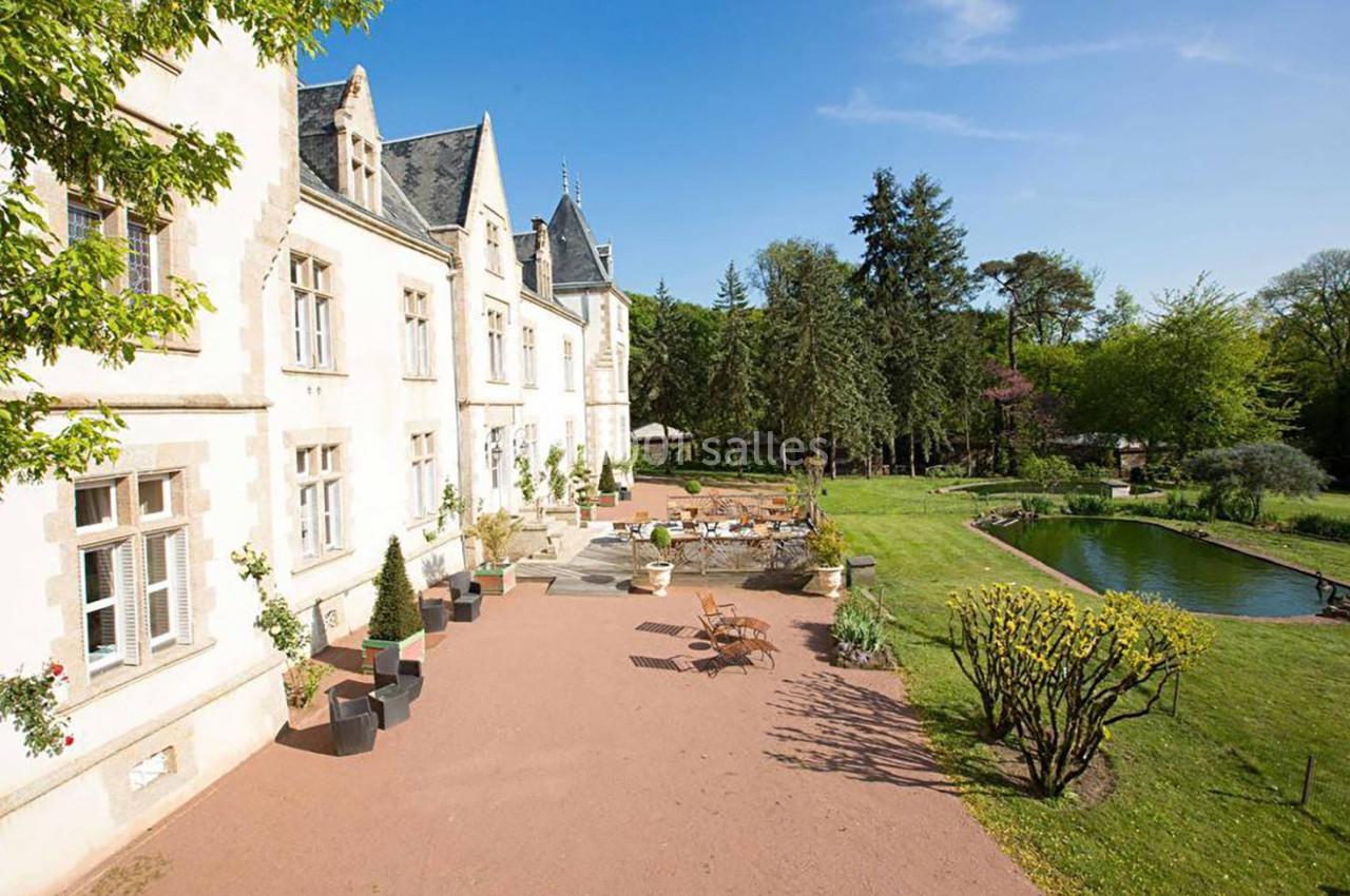 Façade d'un bâtiment historique avec terrasse aménagée, vue sur un jardin arboré et un étang sous un ciel dégagé.