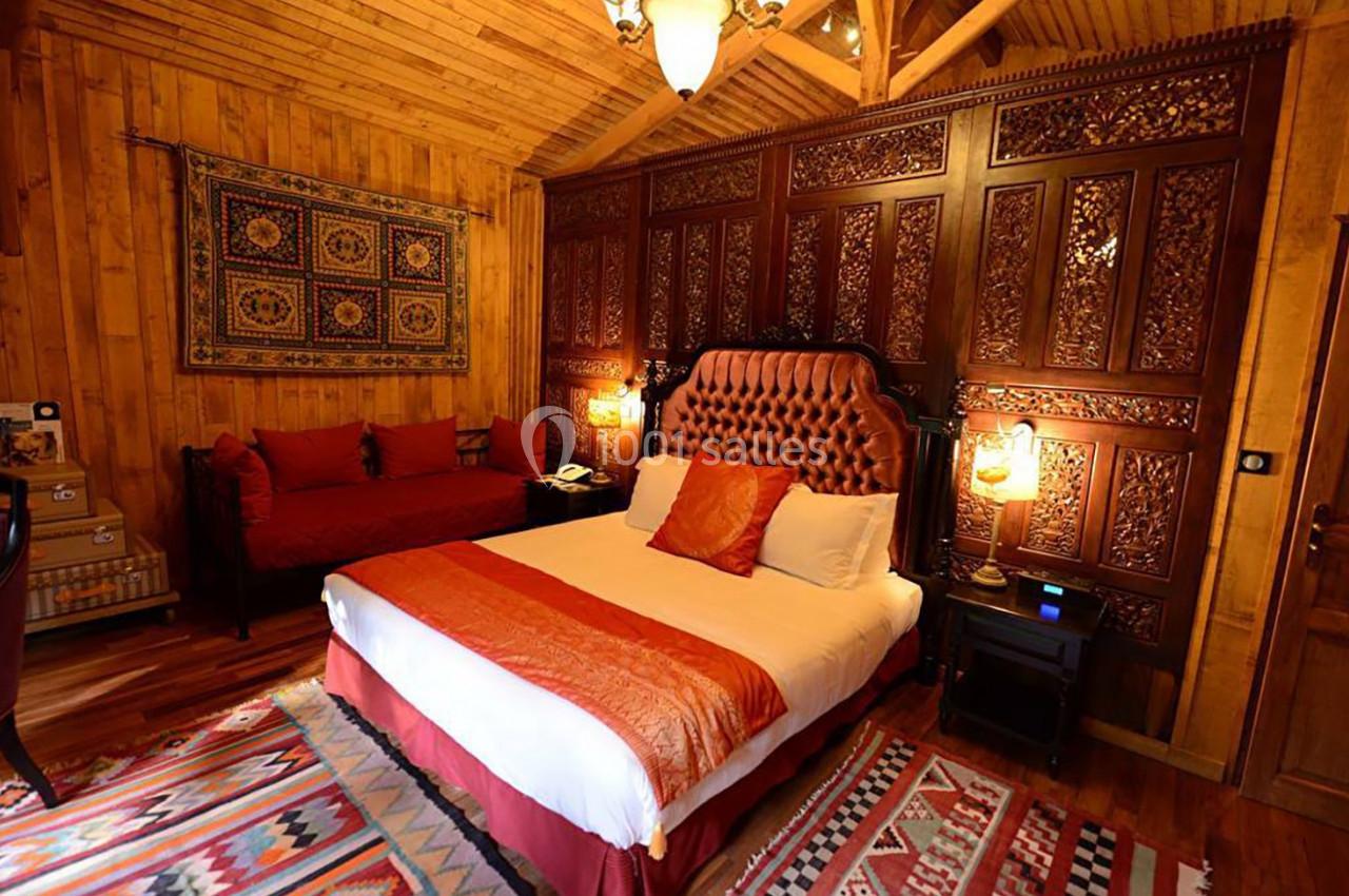 Chambre chaleureuse avec lit double, canapé rouge, murs en bois et panneaux sculptés, éclairée par des lampes douces.