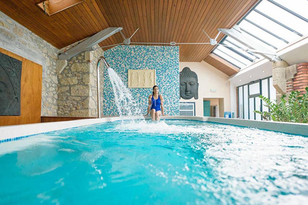 Femme en maillot bleu profitant d'une piscine intérieure avec cascade, entourée de murs en pierre et décorations murales.