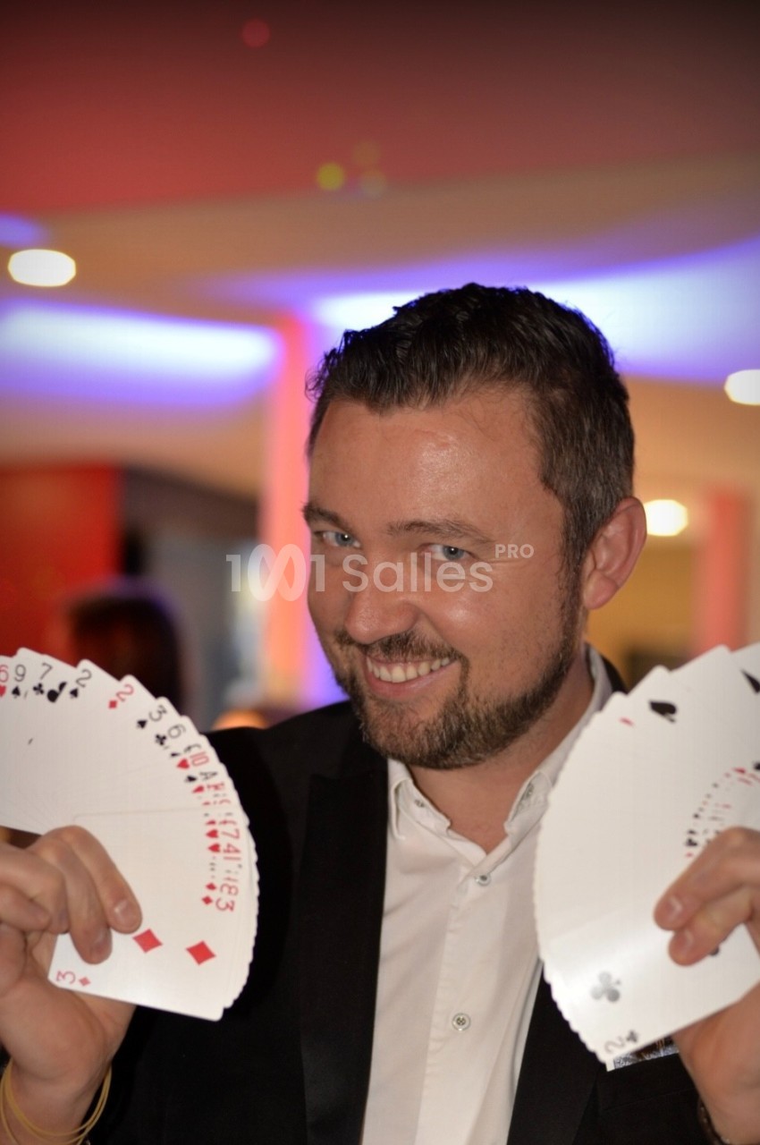 Un homme en costume souriant tient deux éventails de cartes à jouer dans un environnement lumineux.