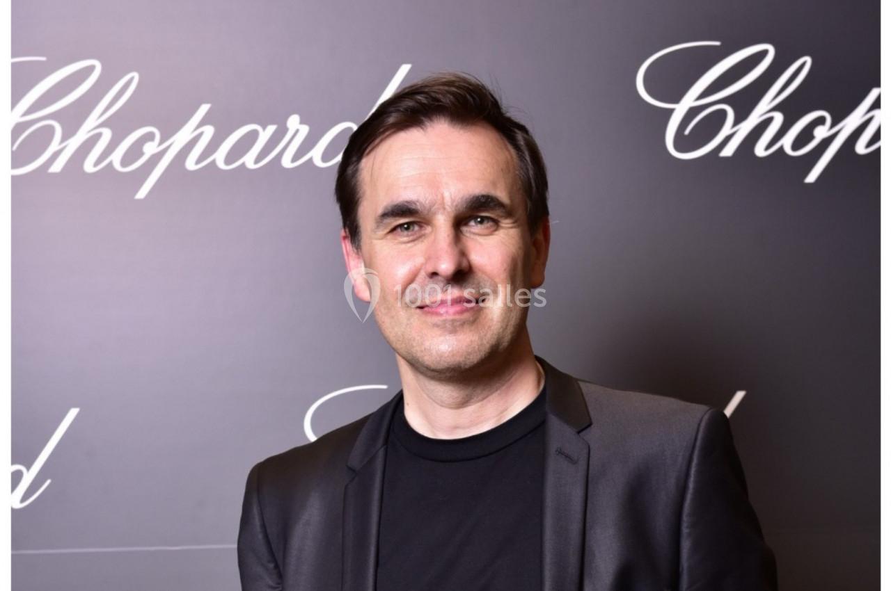 Homme souriant en costume noir devant un fond sombre avec le logo ’Chopard’ répété.