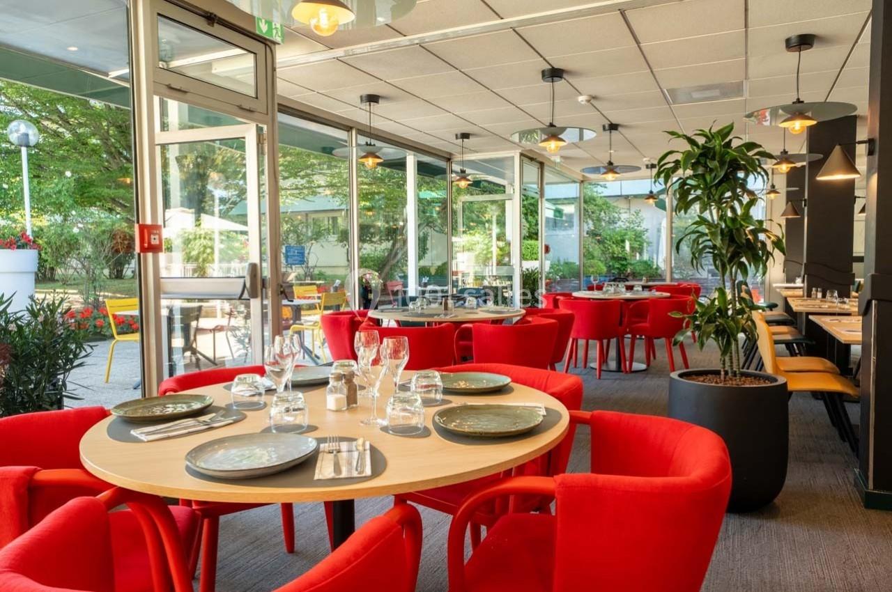 Salle de restaurant lumineuse avec des tables dressées, chaises rouges et vue sur une terrasse arborée.