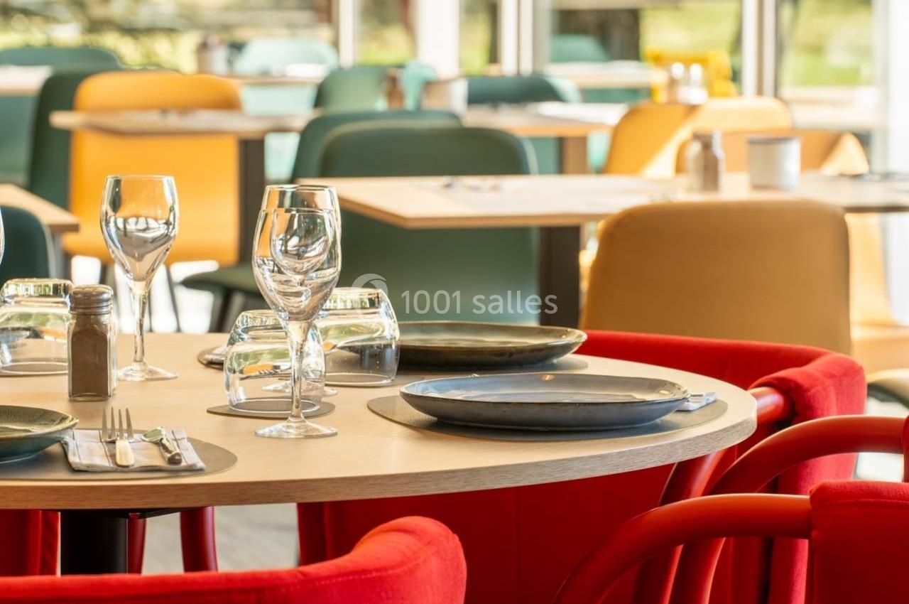 Salle de restaurant lumineuse avec tables dressées, chaises colorées et vue sur l'extérieur.