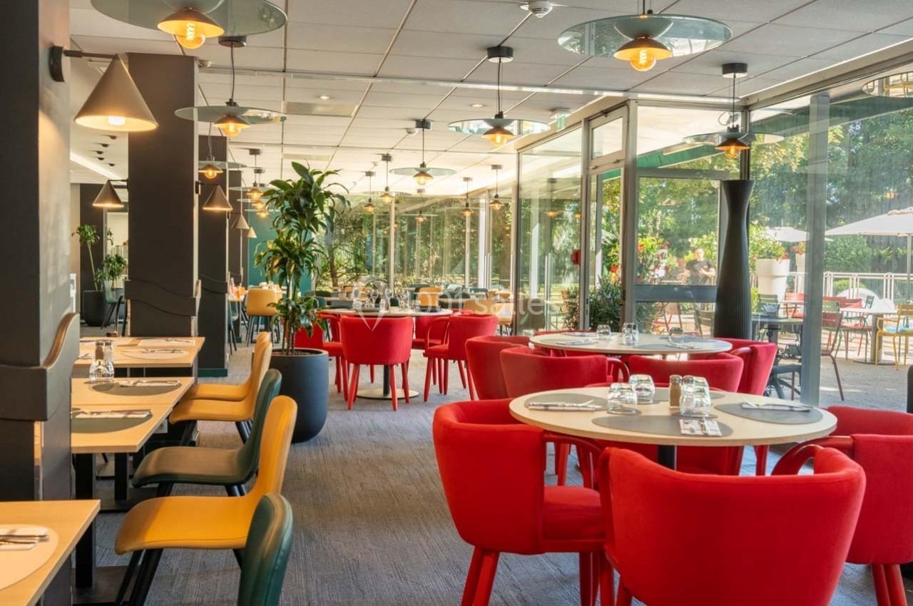 Salle de restaurant lumineuse avec des tables dressées, des chaises colorées et une vue sur un espace extérieur verdoyant.