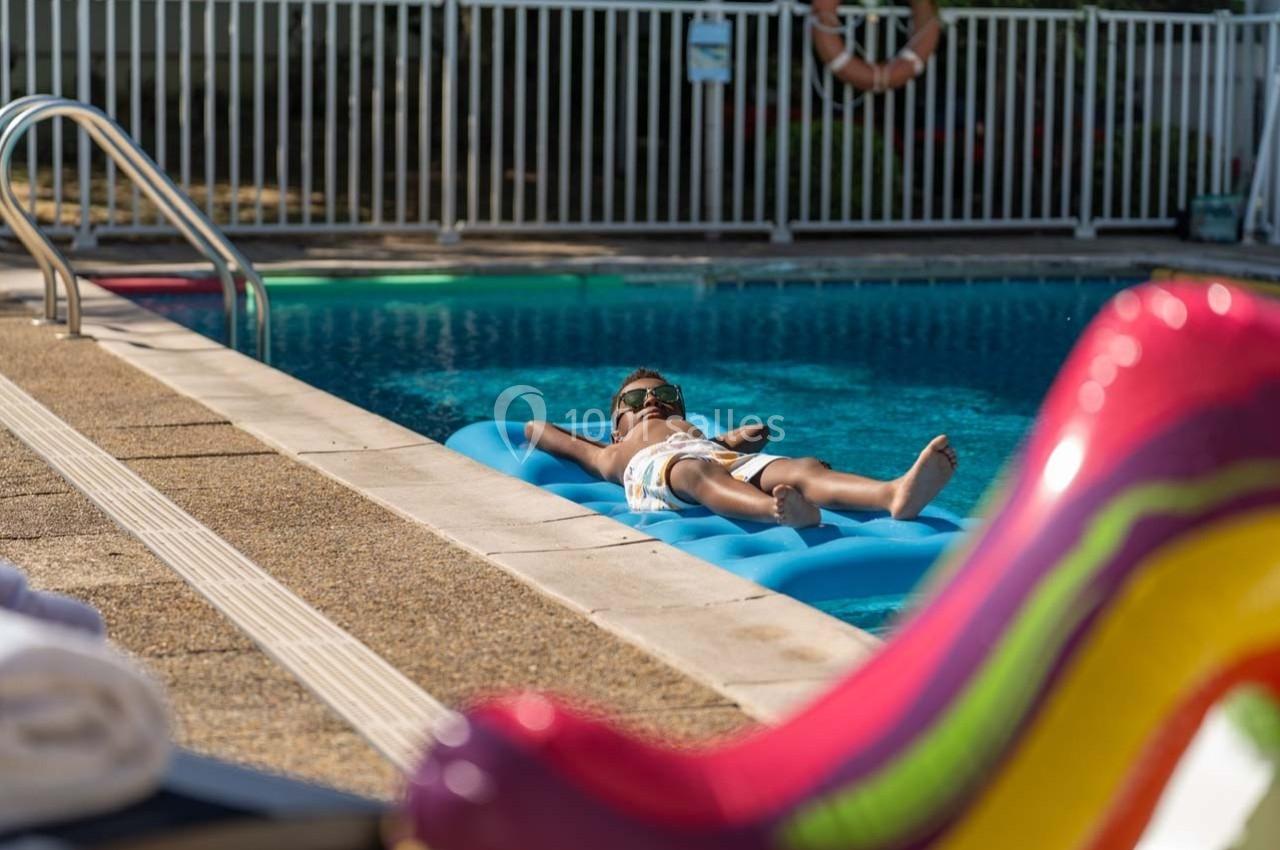 Un enfant allongé sur un matelas gonflable bleu flotte dans une piscine ensoleillée près d'un toboggan coloré.