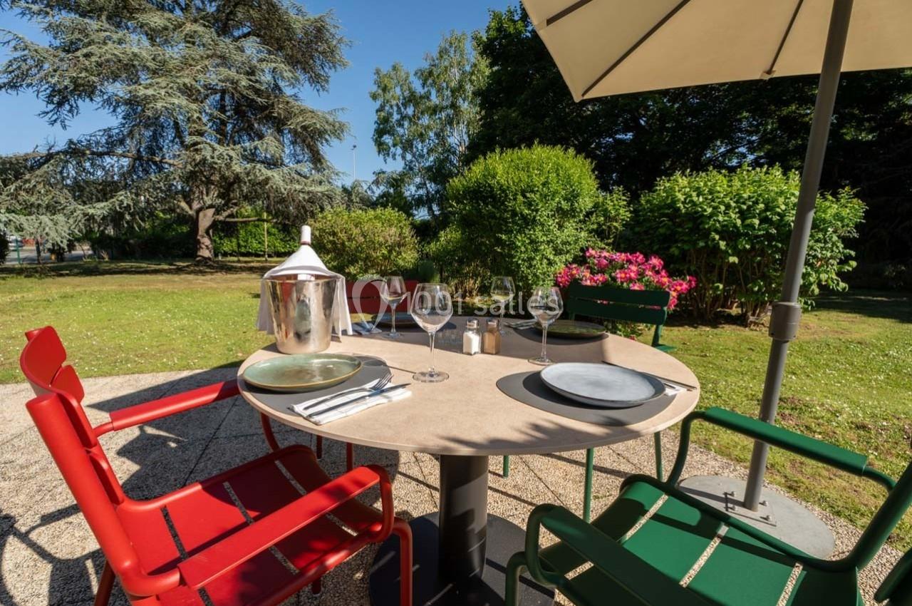 Table de jardin avec chaises colorées, couverts dressés, sous un parasol, entourée de verdure et de fleurs.