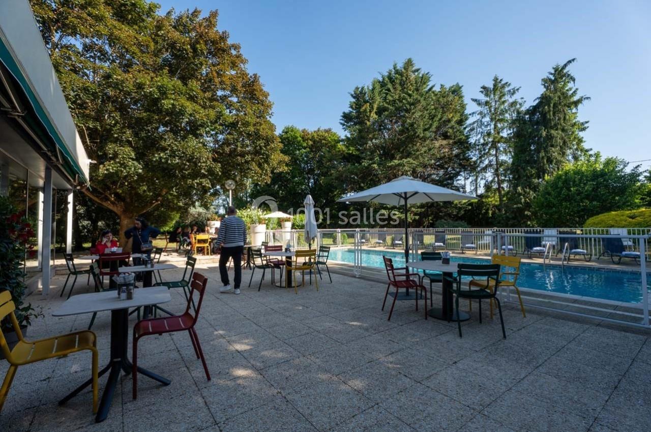 Terrasse avec tables et chaises colorées, bordée d'une piscine et entourée d'arbres sous un ciel dégagé.