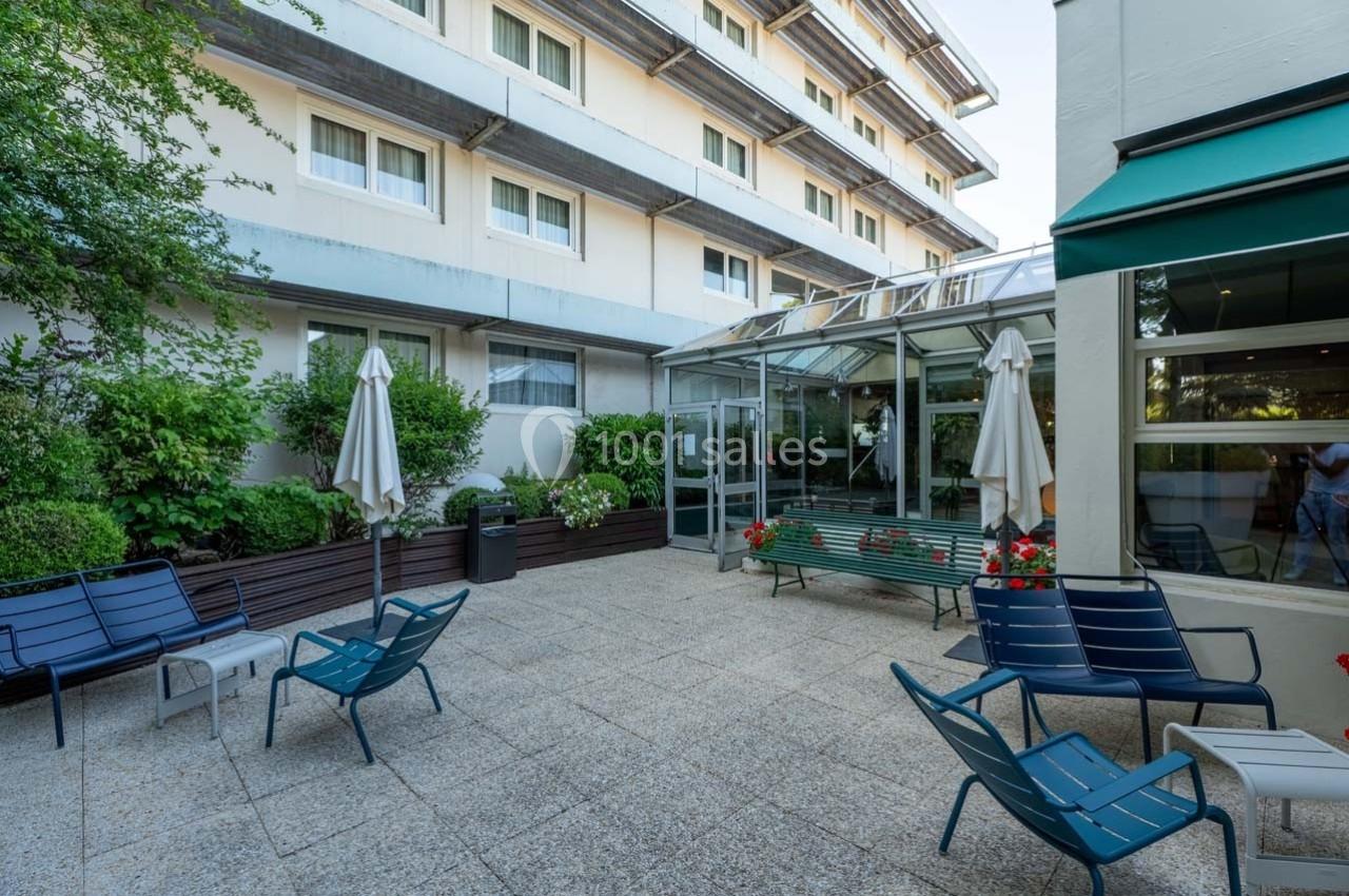 Cour extérieure avec mobilier de jardin, parasols fermés, plantes et accès vitré à un bâtiment moderne.