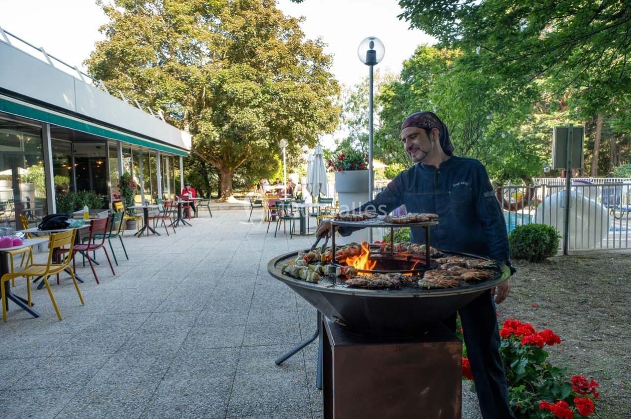 Un cuisinier prépare un barbecue en plein air sur une terrasse entourée de tables et de végétation.