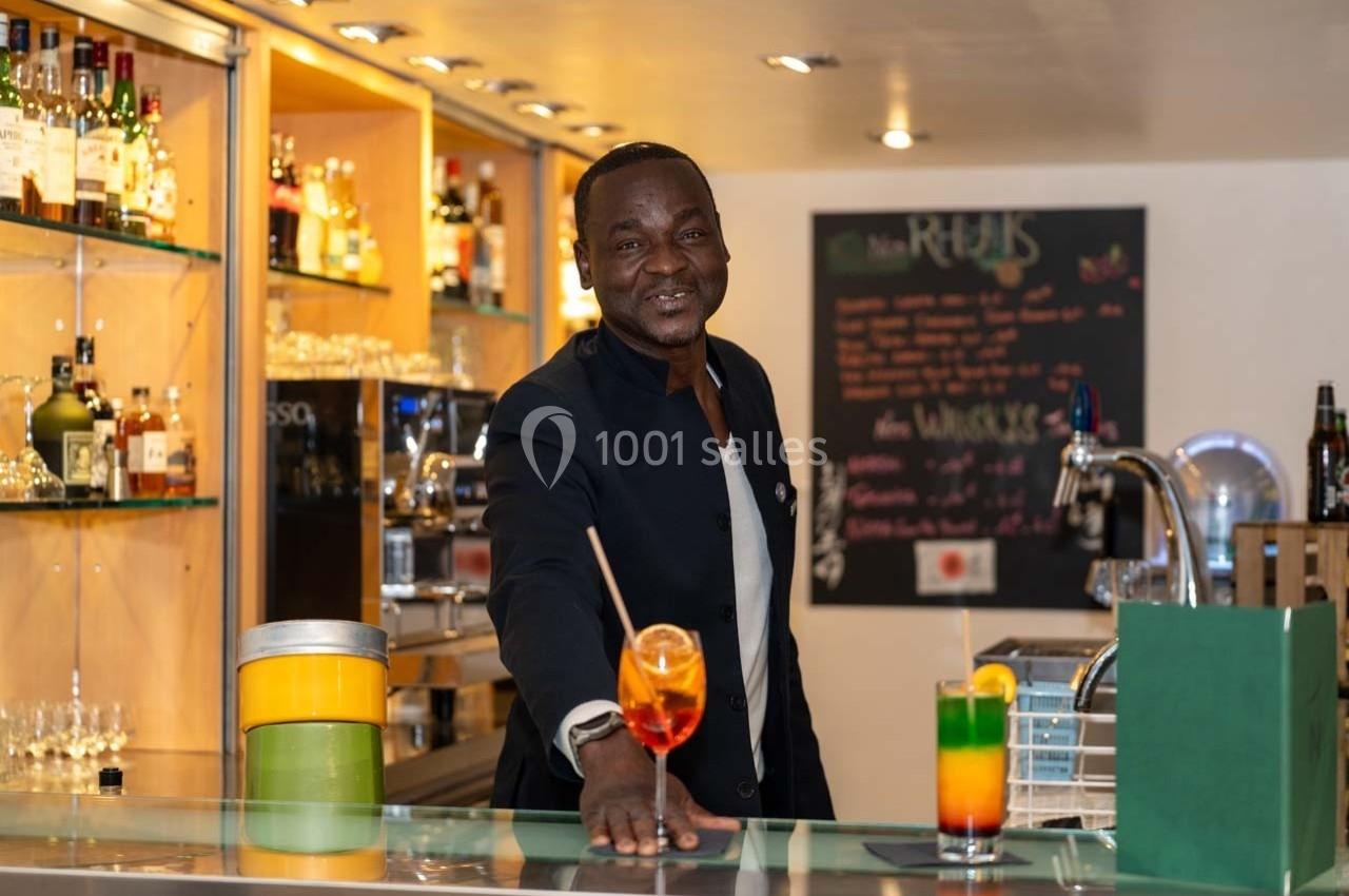 Un barman souriant présente un cocktail coloré sur un comptoir, avec des étagères de bouteilles en arrière-plan.