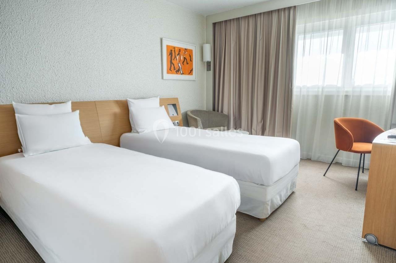 Chambre d'hôtel avec deux lits simples, un bureau, une chaise orange et une grande fenêtre avec rideaux beige.