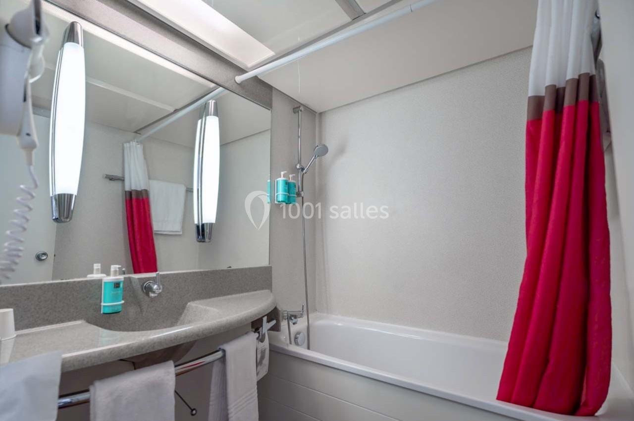 Salle de bain avec lavabo, miroir, baignoire, douchette murale et rideau rouge et blanc.